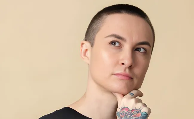 A pensive woman with a buzzcut, arm tattoos and several facial piercings is cupping her chin with one hand
