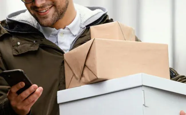 A bearded, smiling man is looking at a smartphone while carrying three packages