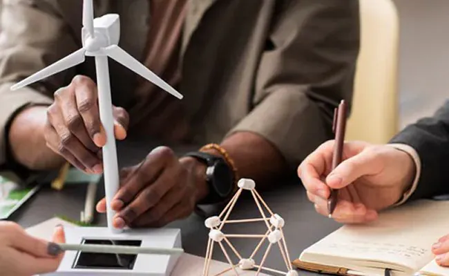 People sitting around a busy desk with one holding a windmill model and the other a notebook and a pen