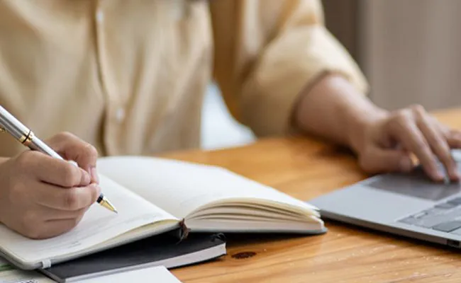 A smiling woman sitting on a desk is working on a laptop while holding a pen over an open notebook