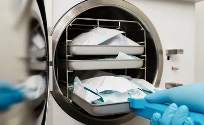 A blue gloved hand is inserting a tray with vacuum sealed bags into an autoclave machine
