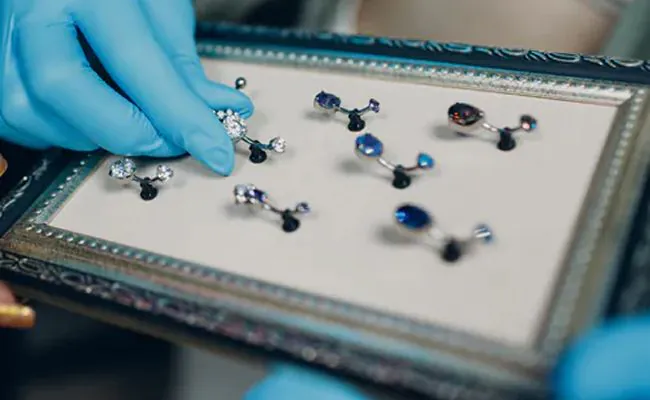 A blue gloved hand is arranging belly button rings placed on a rectangular jewellery display