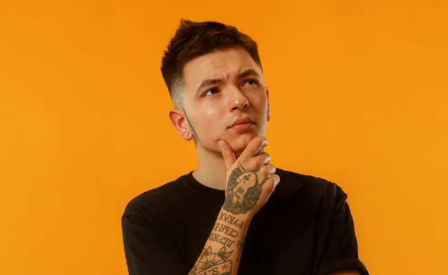 A pensive, tattooed man in a black shirt is holding his chin in one hand