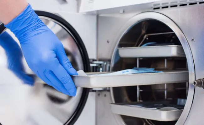 A rubber gloved hand is removing a metal tray from an autoclave machine