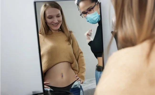 A young woman and her piercer are standing in front of a mirror, inspecting her belly button piercing