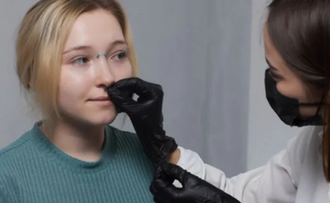 A woman with black gloves and a face mask is holding a piece of nose jewelry to another woman's nose
