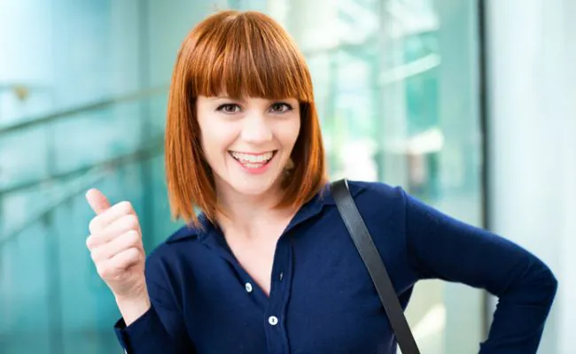 A smiling young woman with red hair and a dark blue blouse is giving a thumbs up
