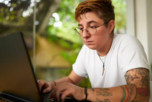 A young person with glasses, several facial piercings and a tattooed arm is working on a laptop.