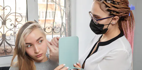 A young woman is using a mirror to inspect a piercing on her ear with her piercer standing next to her