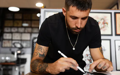 A bearded man with tattoos and a black t-shirt is leaning down on a table, holding a pen.