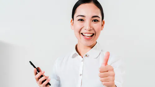 An woman with a white shirt is smiling into the camera holding a phone in one hand and showing a thumbs up with the other