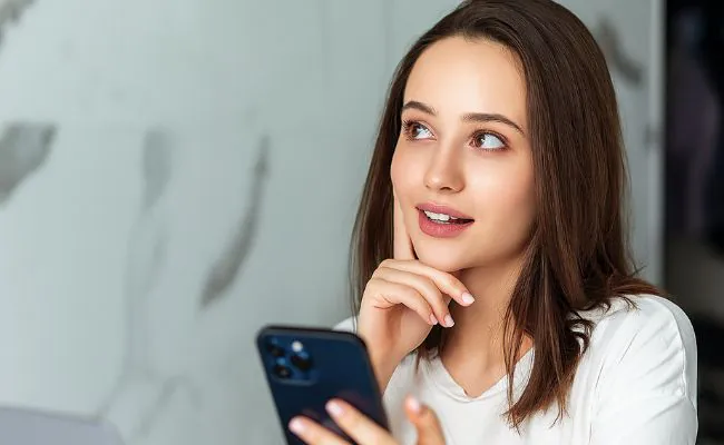 A pensive looking young woman with long brown hair is holding a phone and biting her lip