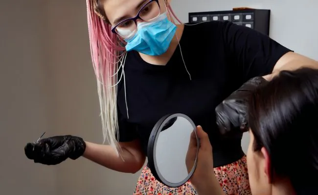 A female piercer wearing a blue face mask and black gloves is looking at a person that is holding up a mirror